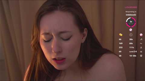 Media: Video of a young Caucasian woman with long brown hair, closed eyes, and slightly open mouth, possibly in a state of distress, set against a blurred, warm-toned background.