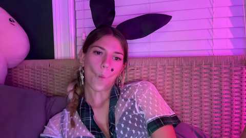 Media: A young woman with light skin, brown hair, and large hoop earrings, wearing a black bunny headband and a plaid shirt, leans against a wicker chair, illuminated by purple lighting.