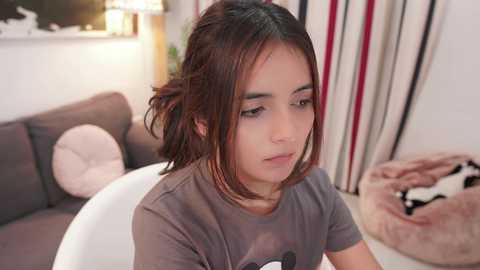 Media: Video of a young woman with medium skin tone and shoulder-length brown hair, wearing a grey t-shirt, sitting on a white chair in a modern living room with a grey sofa, white pillows, and a striped curtain.