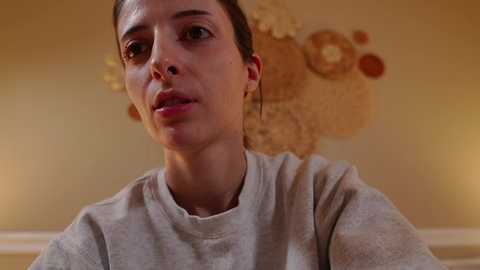 Media: Video of a light-skinned woman with dark hair, wearing a gray t-shirt, looking contemplative. Background features a beige wall with abstract art.