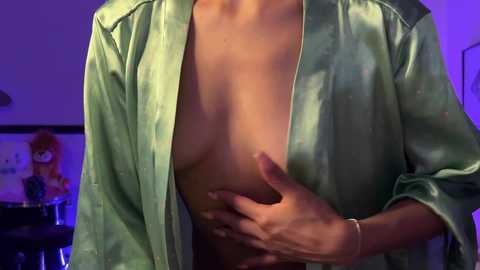 Media: A video of a topless woman with medium breasts, wearing a green satin robe, her hand covering her left breast. Background shows a blurred image of a person with red hair.