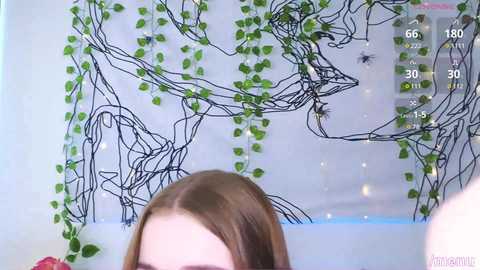 Media: Video of a woman with straight brown hair, partially visible, in front of a minimalist, black line drawing on a white wall with green ivy vines.