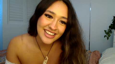 Media: Video of a smiling, light-skinned woman with long brown hair, wearing a beige off-shoulder top and a necklace. Background features a white shuttered window and a potted plant.