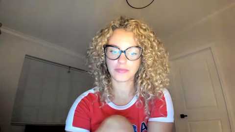 Media: Video of a young woman with curly blonde hair, wearing glasses and a red sports shirt, standing in a dimly lit, white-walled room with a closed door and window blinds in the background.