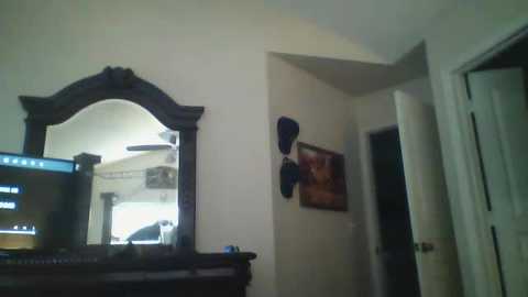 Media: A video of a dimly lit bedroom corner featuring a dark wooden dresser with a mirror, framed photos, and a flat-screen TV. The room has a sloped ceiling and a closed door.