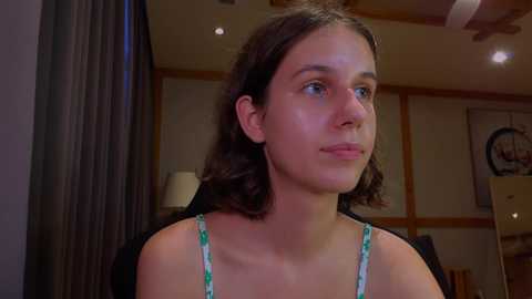 Media: Video of a young Caucasian woman with shoulder-length brown hair, wearing a green floral-patterned spaghetti strap top, looking contemplative in a dimly lit, modern room with wooden accents and a wall clock.