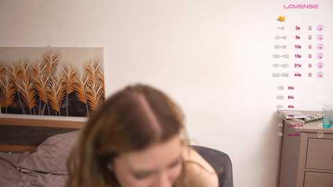 Media: Video of a young woman with shoulder-length brown hair, wearing a beige top, sitting on a bed with a grey pillow, in a room with a \"LOVE\" wall calendar and decorative wheat artwork.