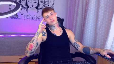 Media: Video of a tattooed, light-skinned man with short, red hair, wearing a black tank top, holding a phone, seated at a desk with a ring light and purple curtains in the background.