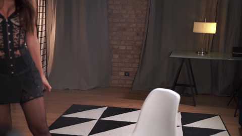 Media: Video of a woman in a black patterned top and shorts, standing beside a modern white chair on a geometric-patterned rug. Background includes a brick wall, a lamp on a black table, and a room divider.