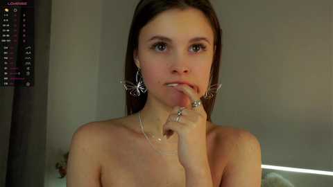 Media: A video of a topless, fair-skinned woman with straight brown hair, wearing butterfly-shaped earrings and a silver necklace, licking her finger in a dimly lit room with a digital display on the left.