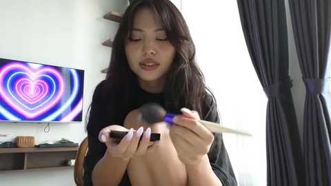 Media: Video of an Asian woman with long black hair, applying makeup on a person's face, holding a makeup brush. Background features a colorful heart-shaped light display and dark curtains.