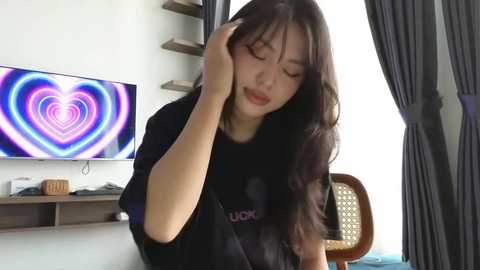 Media: A video of an Asian woman with long black hair, wearing a black shirt, sitting in a modern room with a colorful heart-shaped light on the wall and dark curtains.