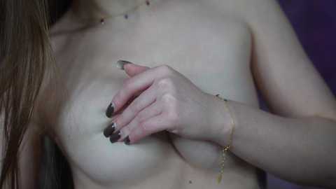 Media: Video of a topless woman with light skin, medium-sized breasts, and long brown hair. She covers one breast with her hand, adorned with black nail polish and a gold bracelet. Background is dark purple.