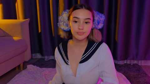 Media: Video of a young Asian woman with light skin, wearing a sailor-style top and floral headband, sitting on a plush rug in a dimly-lit room with purple curtains.