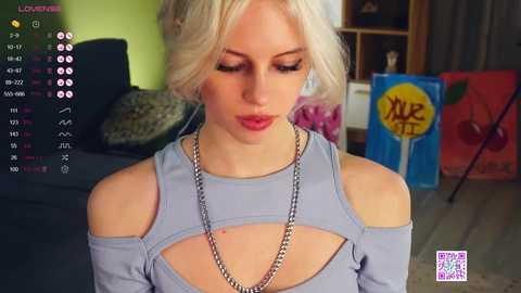 Media: Video of a young blonde woman in a light grey off-shoulder top, with a necklace, indoors, watching live streaming. Background includes colorful artwork and furniture.