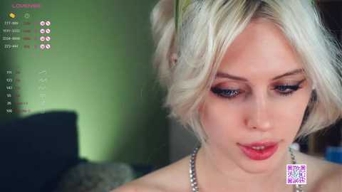 Media: Video of a young, fair-skinned woman with platinum blonde, shoulder-length hair, wearing dark eyeliner and red lipstick, set against a blurred green background.