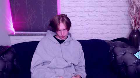 Media: Video of a young, pale-skinned, androgynous person with shoulder-length brown hair, wearing a gray hoodie, sitting on a black leather couch against a white brick wall with purple LED lighting.