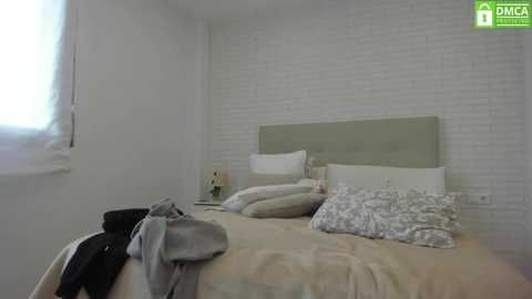 Media: Video of a minimalist bedroom with a white brick wall, a beige bed, and pillows in shades of white and light grey. A small white nightstand with a lamp sits beside the bed.