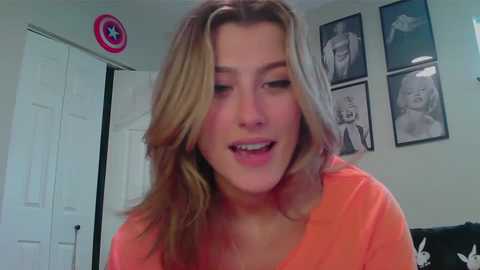 Media: Video of a smiling, light-skinned woman with wavy, shoulder-length blonde hair, wearing an orange shirt, in a room with white doors, a Marvel logo, and framed black-and-white portraits.