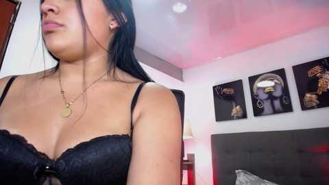 Media: Video of a young Latina woman with long black hair, wearing a black lace bra, standing in a modern bedroom with minimalist decor and three framed paintings on the wall.