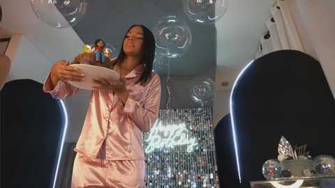 Media: Video of an Asian woman in pink satin pajamas, holding a cake with a figurine, surrounded by floating balloons in a dimly lit, modern room.