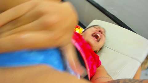 Media: Video of a woman with light skin, wearing a red bikini top and blue shorts, lying on a white leather chair, laughing, background blurred.