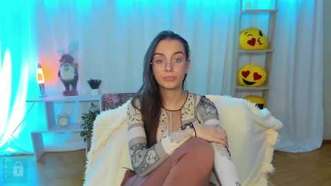 Media: Video of a woman with long dark hair, wearing a striped sweater and pink skirt, sitting on a white chair in a colorful room with a blue light, yellow smiley pillows, and a plant.