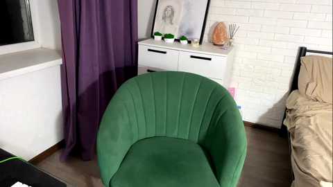 Media: Video of a modern bedroom featuring a green, curved armchair, a white dresser with artwork and green plants, a bed with beige bedding, and a purple curtain.