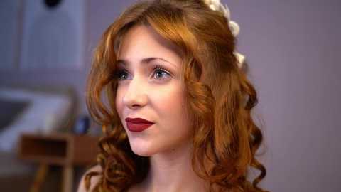 Media: Video of a young Caucasian woman with wavy, auburn hair and fair skin, wearing red lipstick, indoors with blurred furniture in the background.