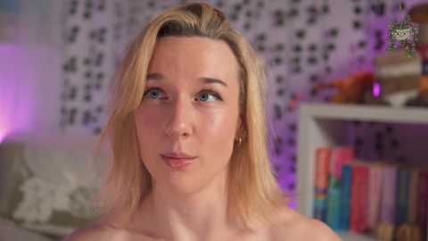 Media: Video of a fair-skinned, blonde woman with blue eyes, wearing minimal makeup, in a cozy room with a patterned wall, bookshelves, and a soft purple glow.