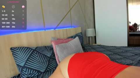 Media: Video of a woman in a red tank top lying on a bed with blue geometric-patterned sheets and pillows. The background includes a wooden headboard and a modern room with a visible TV displaying a game interface.