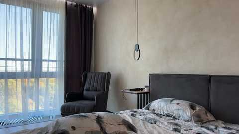 Media: A video of a modern, minimalist bedroom featuring a gray upholstered bed, a gray armchair, sheer white curtains, and a black and white patterned bedspread.