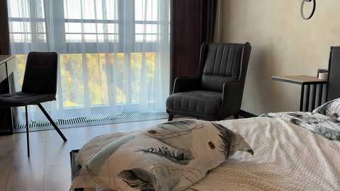 Media: Video of a modern, minimalist bedroom with a gray upholstered chair, white bed with a quilt, a wooden desk, and a black chair by a window with sheer white curtains.