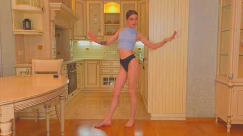 Media: Video of a slender, fair-skinned woman with dark hair in a light blue crop top and black panties, dancing barefoot in a warm, wooden kitchen.