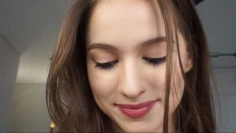 Media: Video of a young woman with fair skin, long brown hair, and closed eyes, wearing subtle makeup with pink lipstick and black eyeliner, smiling softly.