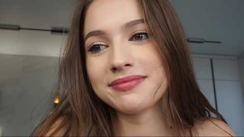 Media: Video of a young woman with long brown hair, fair skin, and light makeup, smiling slightly. She is indoors with a blurred background featuring white walls and a ceiling light.