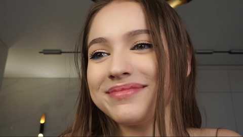 Media: Video of a young woman with light skin and long brown hair, smiling, wearing a black top. Background features a modern interior with a lamp and white walls.