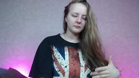 Media: Video of a young Caucasian woman with long, straight brown hair, wearing a black sweatshirt with a distressed Union Jack design, smiling, holding her hair, against a textured light purple background.