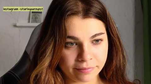 Media: Video of a young woman with fair skin, brown hair, and light brown eyes, wearing a black shirt, in a home setting with a white wall and a small framed picture.