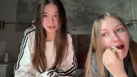 Media: Video of two young women with long brown hair, one wearing a white Adidas jacket, the other in a grey jacket, sitting on a bed in a dimly lit room with floral wallpaper.