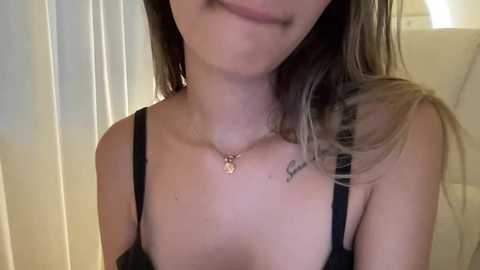 Media: Video of a young woman with fair skin, brown hair, and a slender build, wearing a black bra, with a necklace featuring a pendant, indoors.