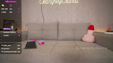 Media: Video of a cozy, plush living room with a beige sectional sofa, featuring a large, realistic, red and pink dildo plush toy, and a laptop open on the couch.