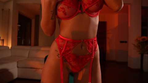 Media: Video of a woman in red lace lingerie, garter belt, and stockings, posing indoors with dim lighting. Her medium-sized breasts are partially visible. Background features a white couch and a potted plant.