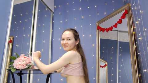 Media: Video of a young Caucasian woman with long brown hair, wearing a pink crop top, smiling and posing in a room adorned with blue walls, heart decorations, and floral arrangements.