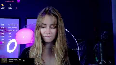 Media: Video of a young blonde woman with long hair, wearing a black top, looking down with a slight smile, in a dimly lit room with a pink balloon, computer screens, and a lamp.