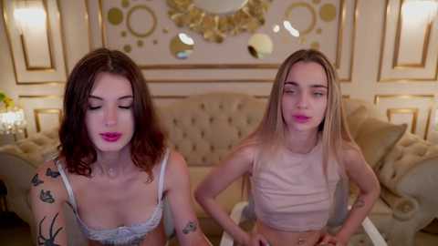 Media: Video of two young women with fair skin, one with curly brown hair and the other with straight blonde hair, wearing light-colored crop tops, posing in a stylishly decorated living room with gold accents and plush furniture.