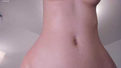 Media: Video of a nude, fair-skinned, slender woman's torso from waist to chest, showing a smooth, hairless body with a small navel. The background is a neutral, softly lit indoor space with white walls and a ceiling.