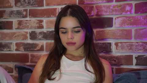 Media: Video of a young woman with long, straight brown hair, wearing a white tank top, against a rustic brick wall with purple lighting.