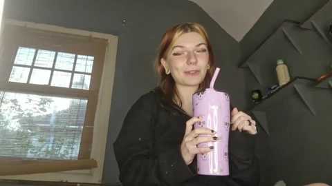 Media: Video of a smiling woman with light skin and auburn hair, wearing a black hoodie, holding a pink bubble tea cup. The room has gray walls, a window with blinds, and a gray shelf with items.