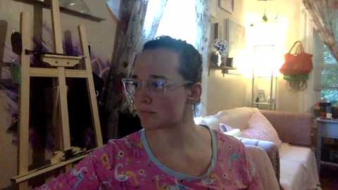 Media: Video of a woman with glasses, wearing pink pajamas with cartoon characters, sitting in a cozy, dimly lit living room with a white couch, art supplies, and a window with sheer curtains.